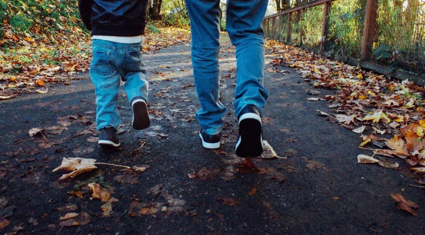 An adult and a small child in jeans walking side by side down a path covered in autumn leaves, seen from behind