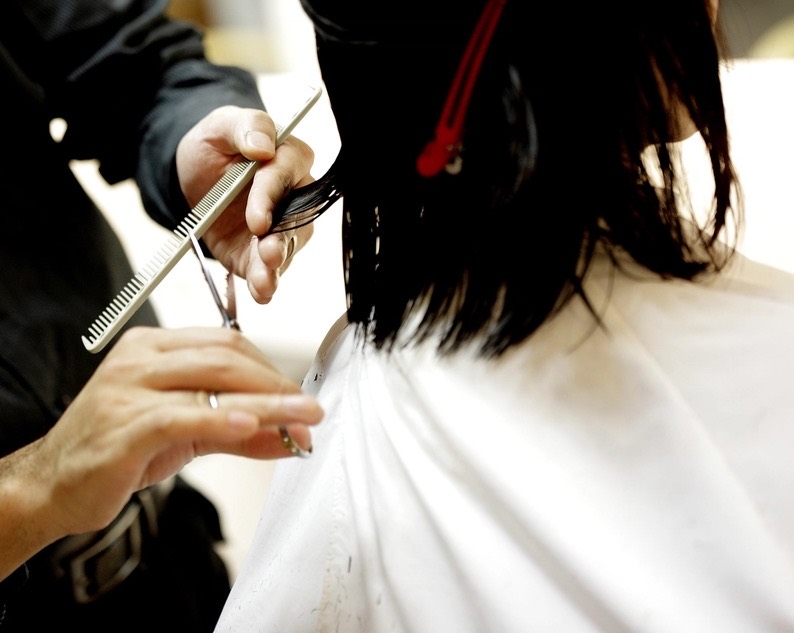 A stylist's hands holding a comb and shears, trimming a client's dark hair from behind