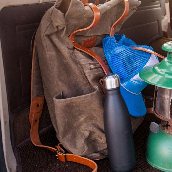 A waxed canvas rucksack with leather straps beside a water bottle and camp lantern