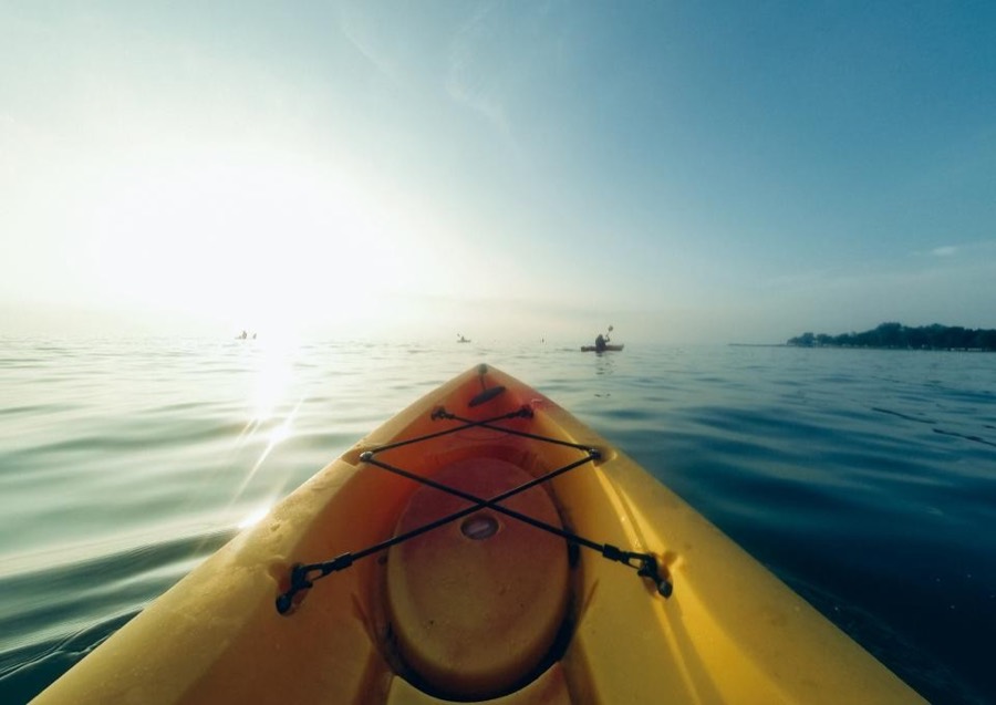 Looking over the yellow bow of a kayak toward other paddlers on still, sunlit water