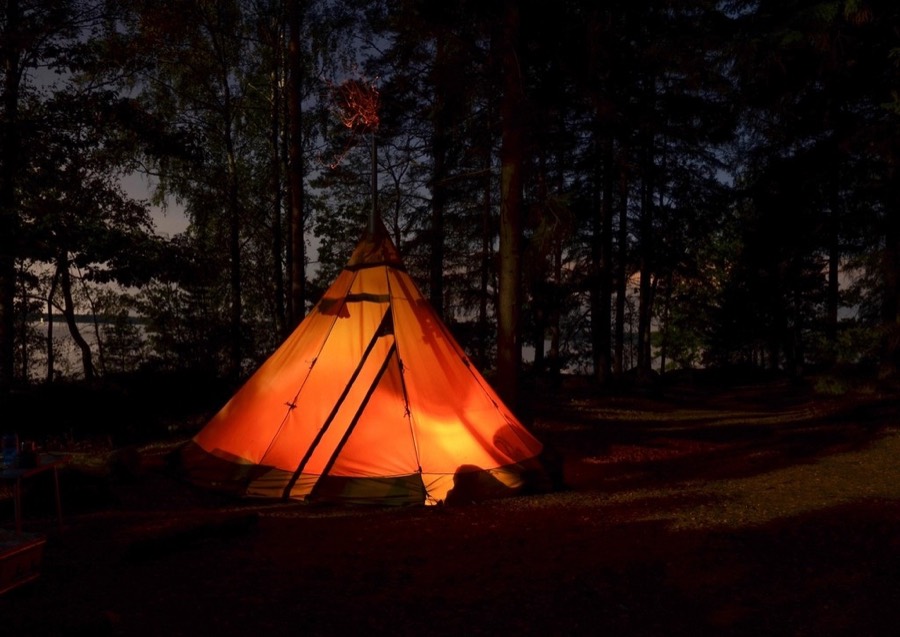 A canvas tipi glowing orange from a lantern inside, pitched among dark trees at night