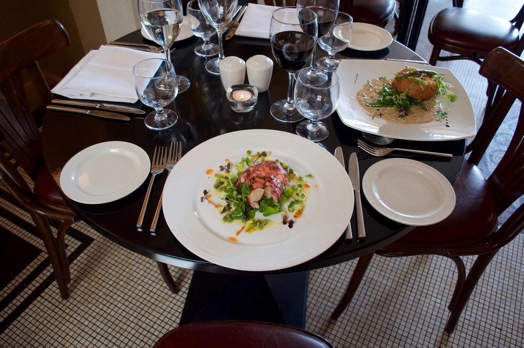 A round dark table set with white plates, wine glasses and a plated first course, on a small-tile floor