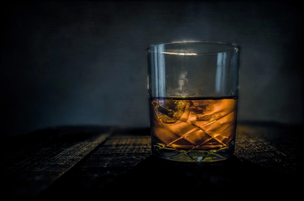 A cut-crystal rocks glass of bourbon with ice on a dark wooden bar