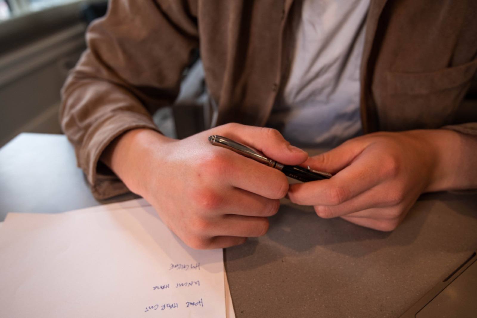 Two hands holding a silver pen over a sheet of handwritten notes on a grey table