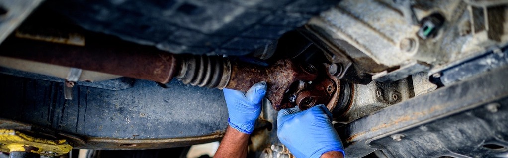Gloved hands working on a rusted driveshaft joint under a vehicle raised on a shop lift