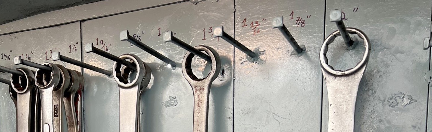 Heavy box-end wrenches hanging on pegs, each size painted by hand on the steel wall above it