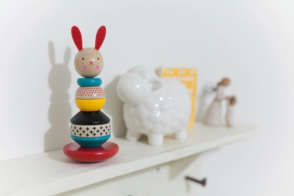 A wooden stacking toy in red, yellow and teal on a white shelf beside a ceramic lamb