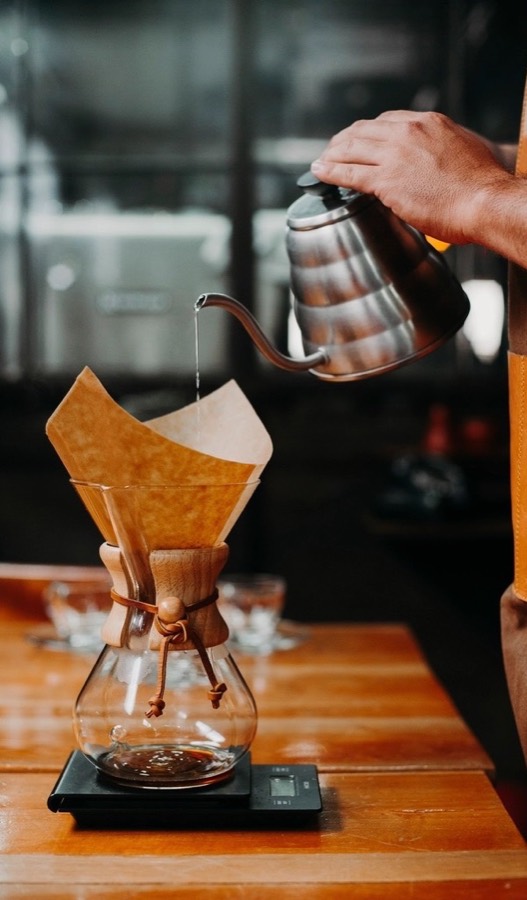 A hand pouring hot water from a gooseneck kettle into a paper filter on a glass brewer, set on a scale