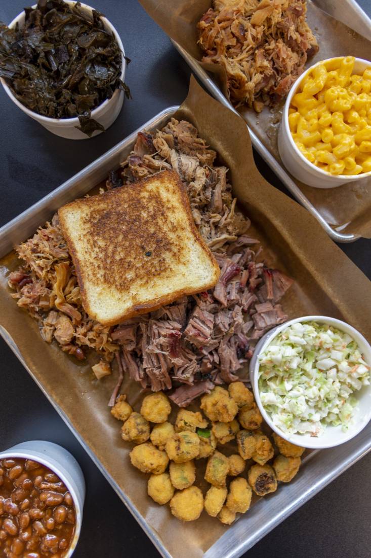 A sheet pan lined with butcher paper, piled with pulled pork and chopped brisket, a slice of thick toast, fried okra, slaw, mac and cheese and beans on the side