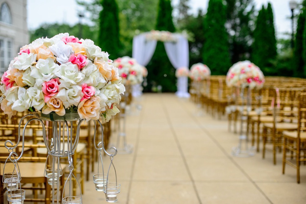 Rows of wooden ceremony chairs lining an aisle toward a draped arch, with flower stands along the edge