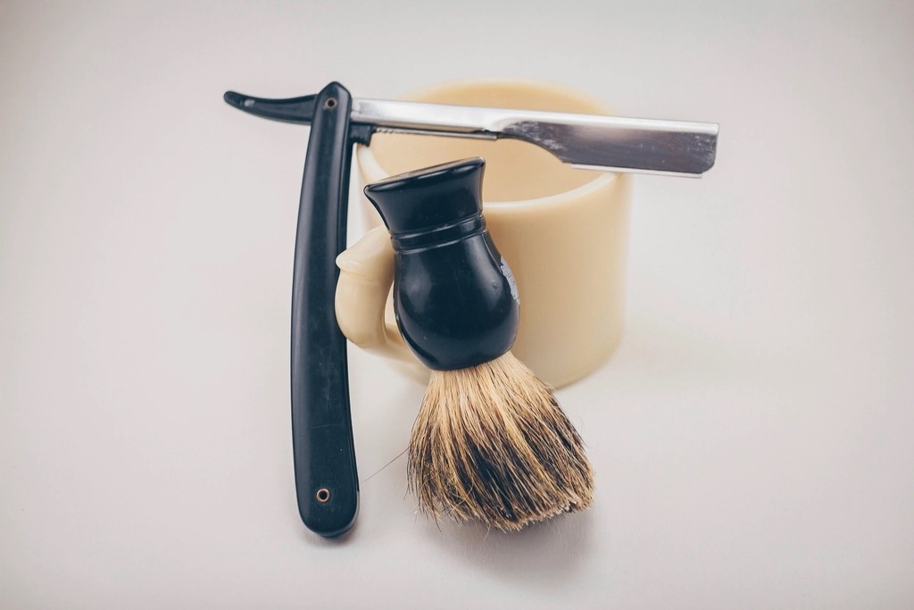 A straight razor with a black handle, a badger shaving brush and a cream shaving mug on a pale table