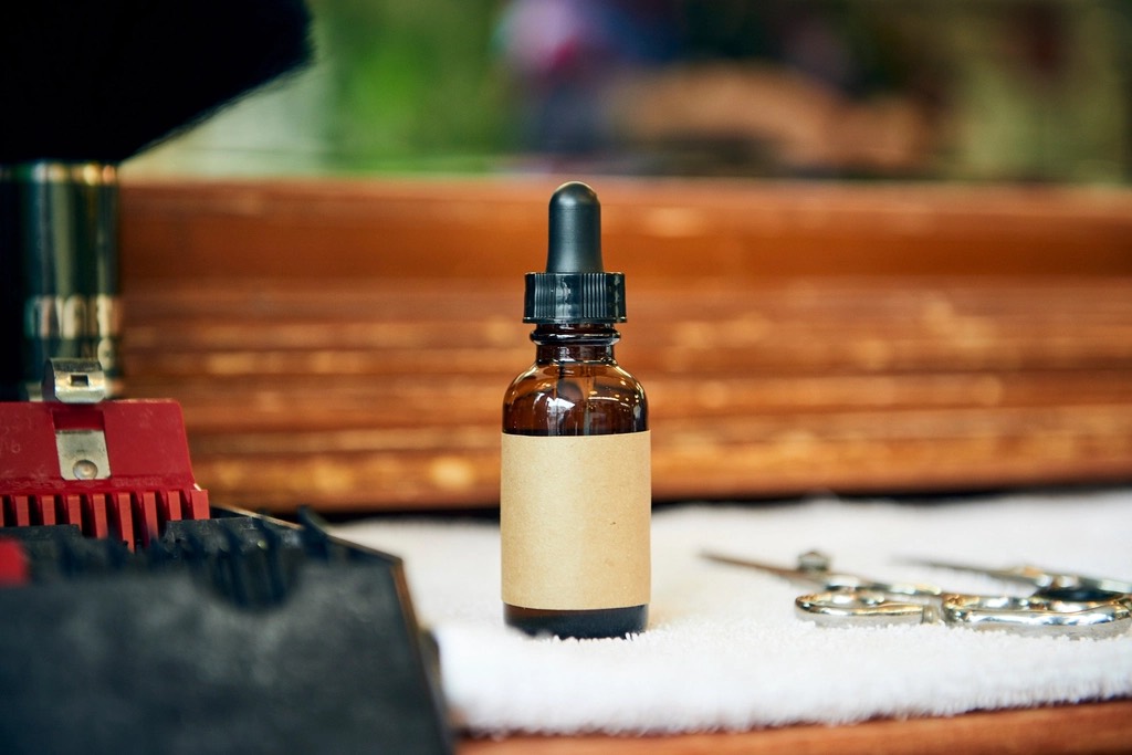 A small amber dropper bottle of beard oil with a plain kraft label on a white towel, scissors beside it