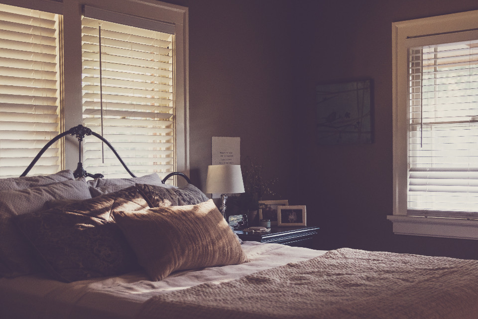 A made bed with an iron headboard and linen pillows in a corner room, morning light through blinds on two walls