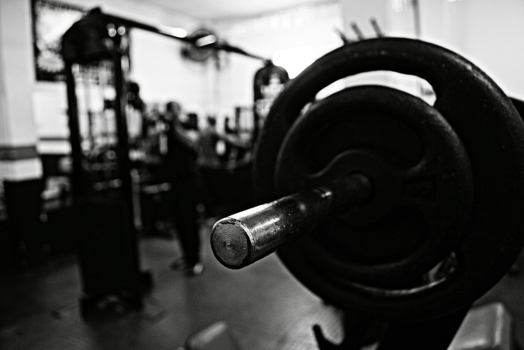 Black and white close-up of a loaded barbell sleeve, with squat racks blurred in the background