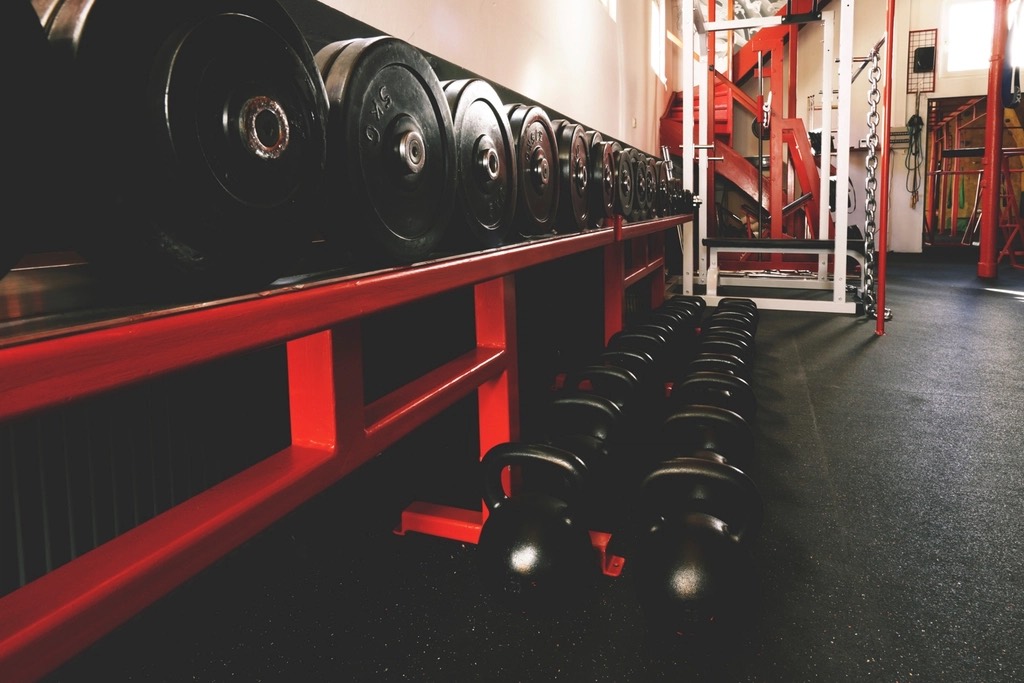 A long rack of bumper plates above two rows of kettlebells on a rubber floor, a squat rack beyond