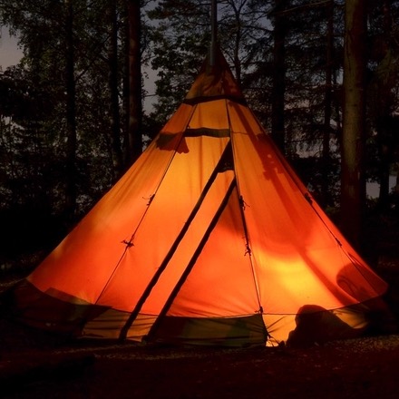 An orange canvas tipi tent lit from inside at dusk among trees