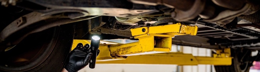 The underside of a car raised on yellow lift arms, a gloved hand shining a flashlight up at it