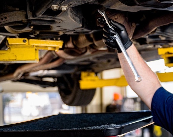 A gloved hand loosening a drain plug under a car on a lift, a drain pan below