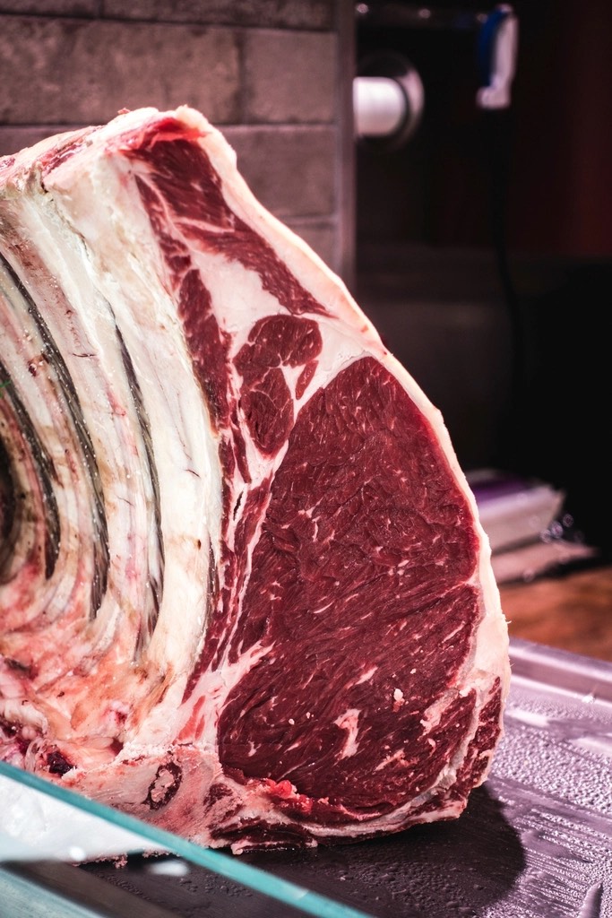 A bone-in beef rib section with deep red marbled eye, resting on a glass shelf in a chilled case
