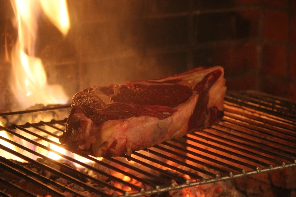A thick bone-in steak searing on a steel grate over open oak flames