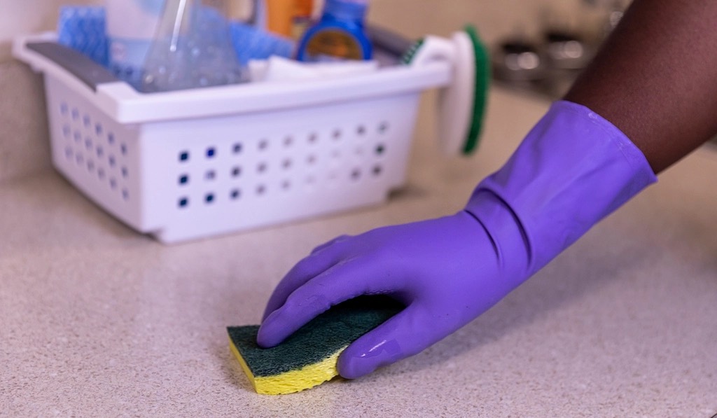 A gloved hand scrubbing a speckled kitchen counter with a sponge, a basket of cleaning bottles behind it