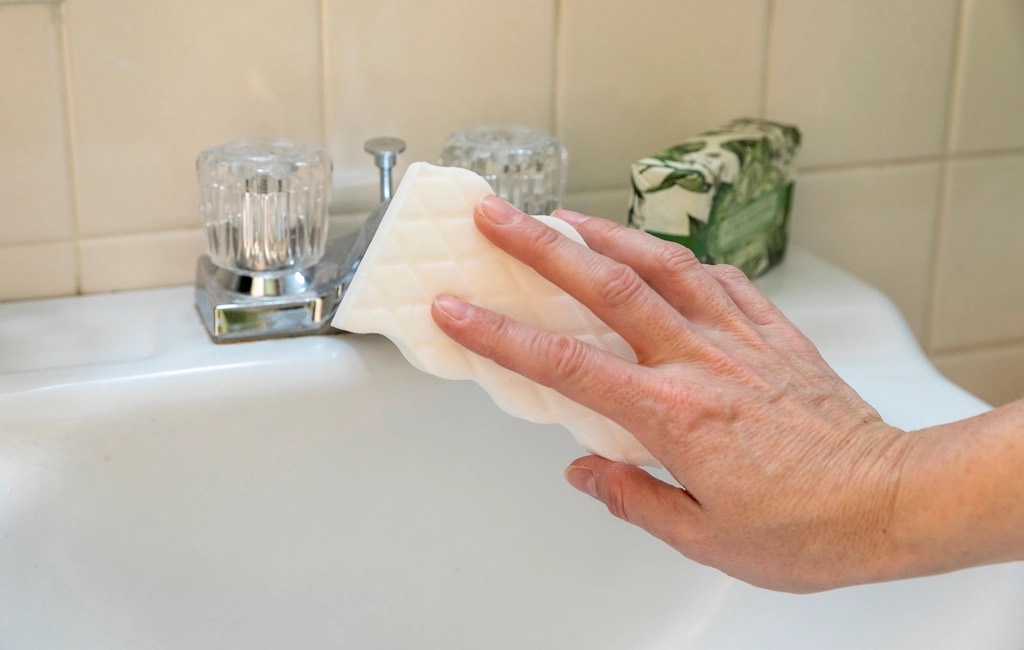 A hand wiping the base of a chrome faucet on a white bathroom sink