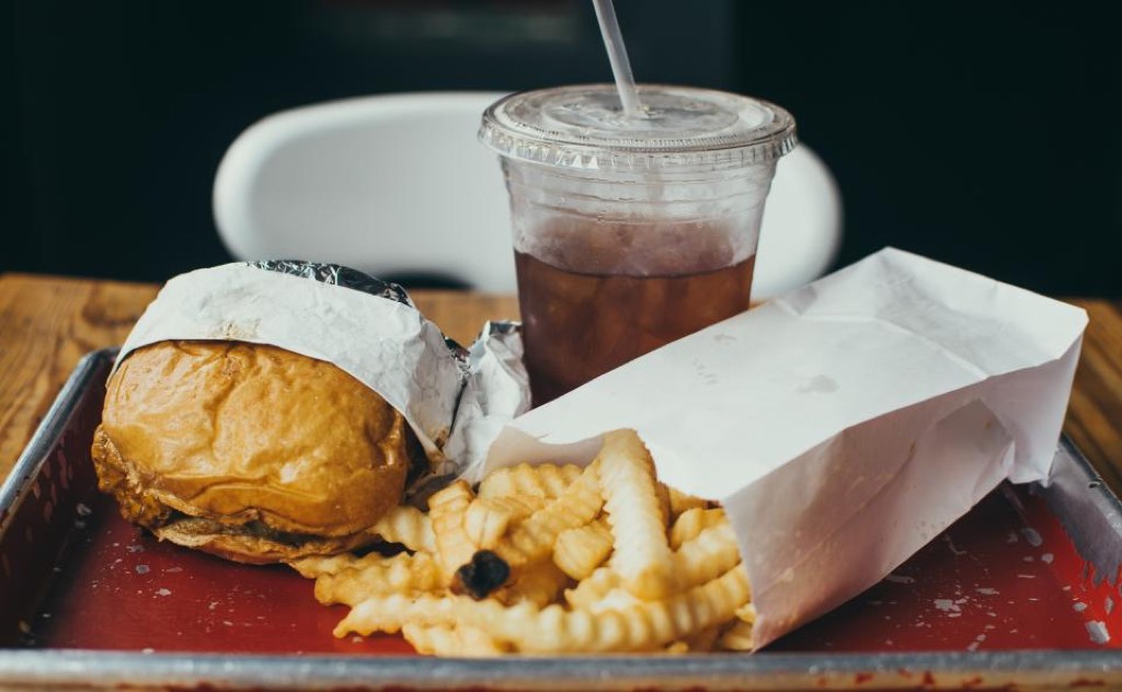 A burger half-wrapped in paper beside a bag of crinkle fries and an iced drink on a red metal tray