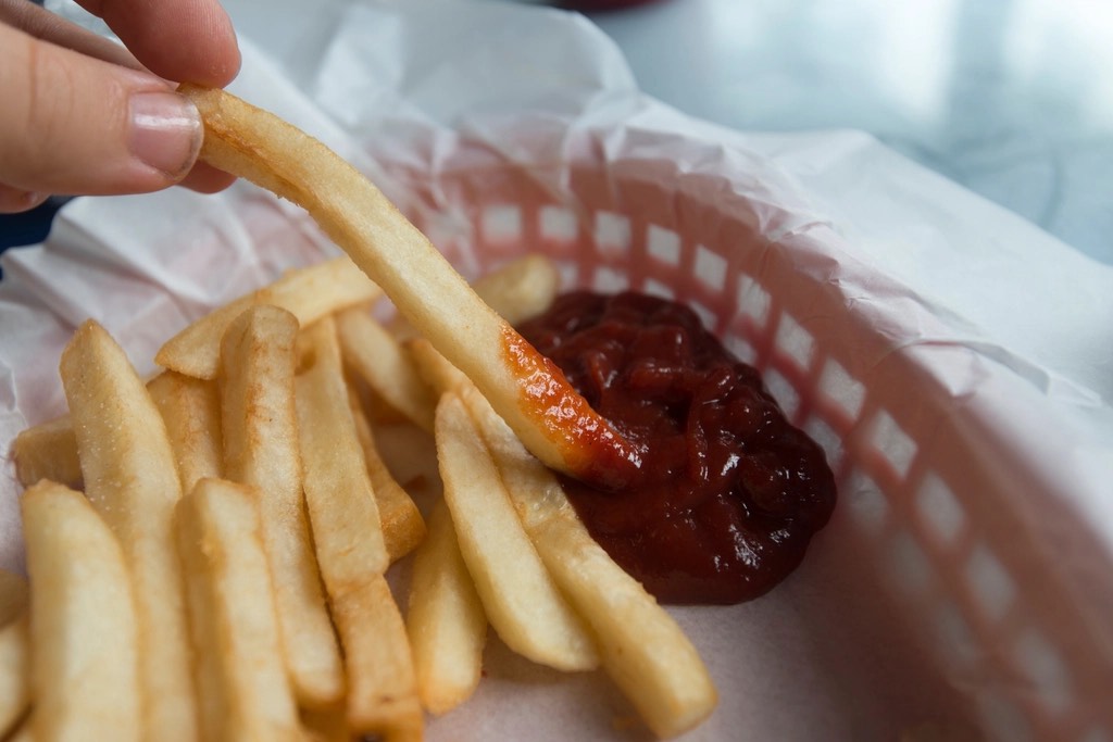 A hand dipping a golden French fry into ketchup in a paper-lined red basket of fries