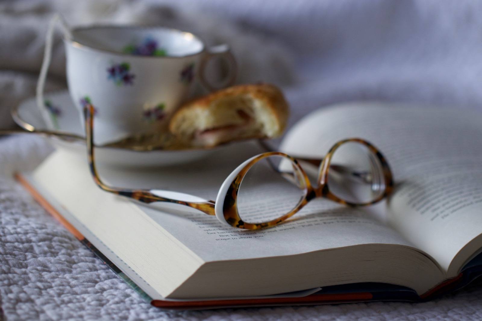 Tortoiseshell reading glasses resting on an open book beside a flowered teacup and a pastry