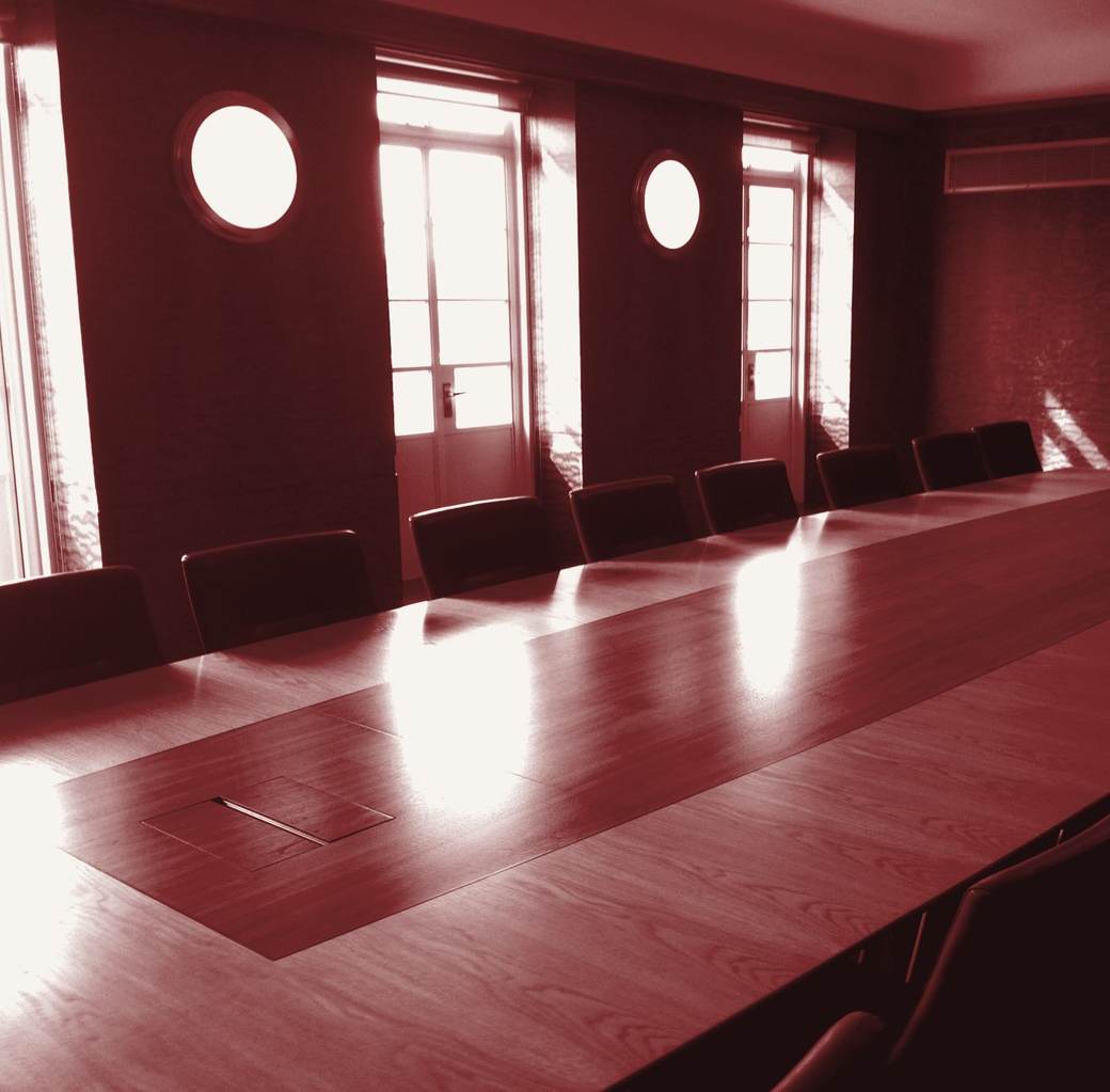 An empty conference room with a long polished table, leather chairs and tall windows letting in afternoon light