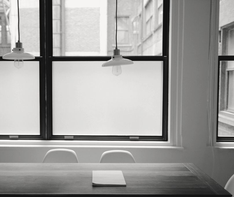A plain wooden table with a single notepad under tall windows that look out on old brick buildings