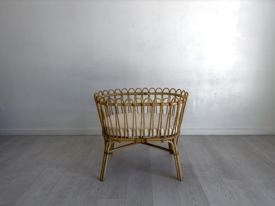 A woven rattan bassinet with a cream mattress standing in an empty white room with a pale wooden floor