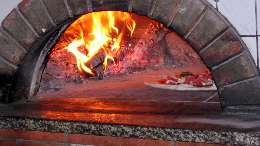 The mouth of a brick wood-fired oven, oak logs burning at the back and a pizza baking on the deck near the opening