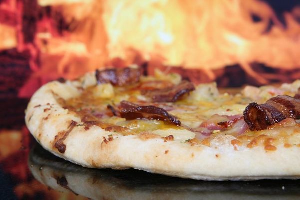 A pizza with bacon and melted cheese on the oven deck, flames glowing behind it