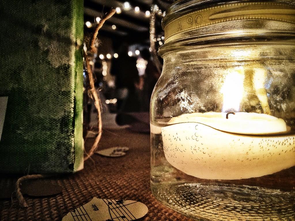 A candle floating in a glass jar on a burlap runner, with string lights blurred along the barn beams behind it