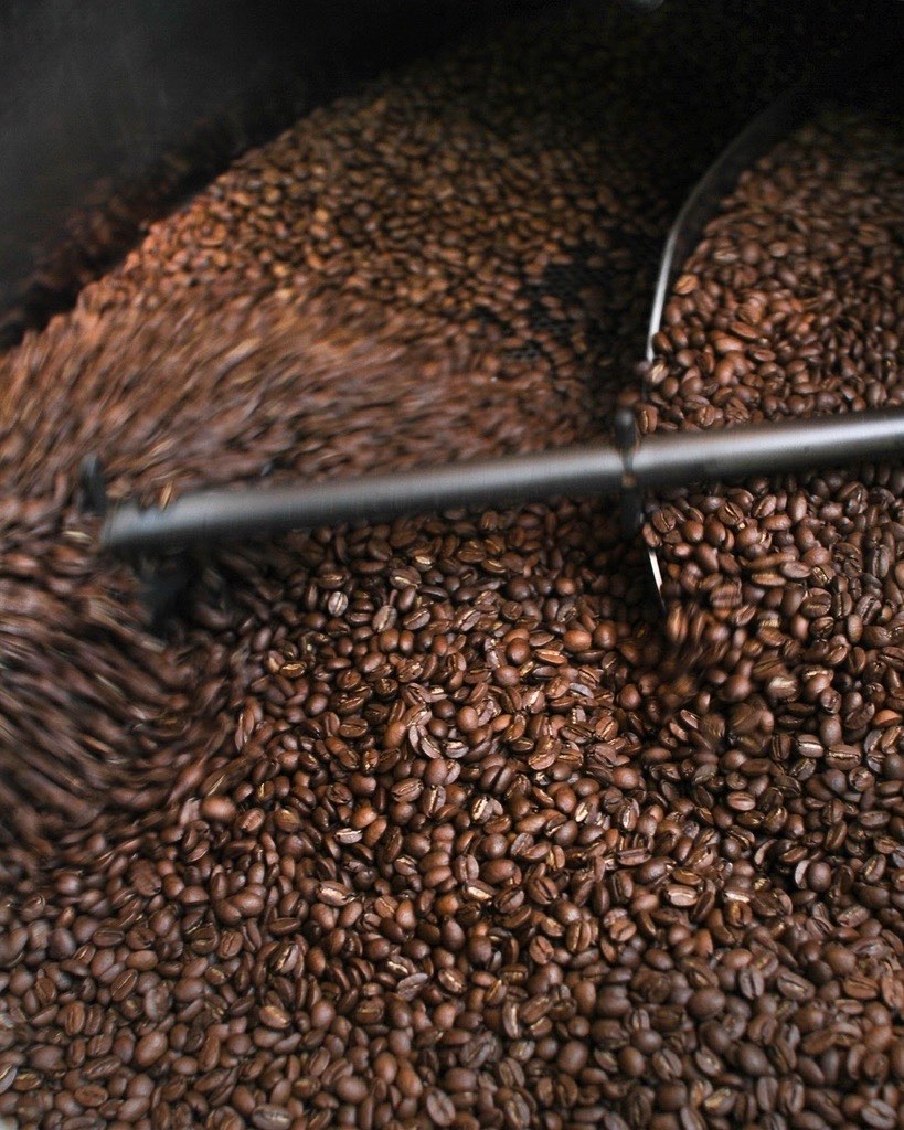 Freshly roasted coffee beans turning in a roaster's cooling tray, a steel stirring arm sweeping through them
