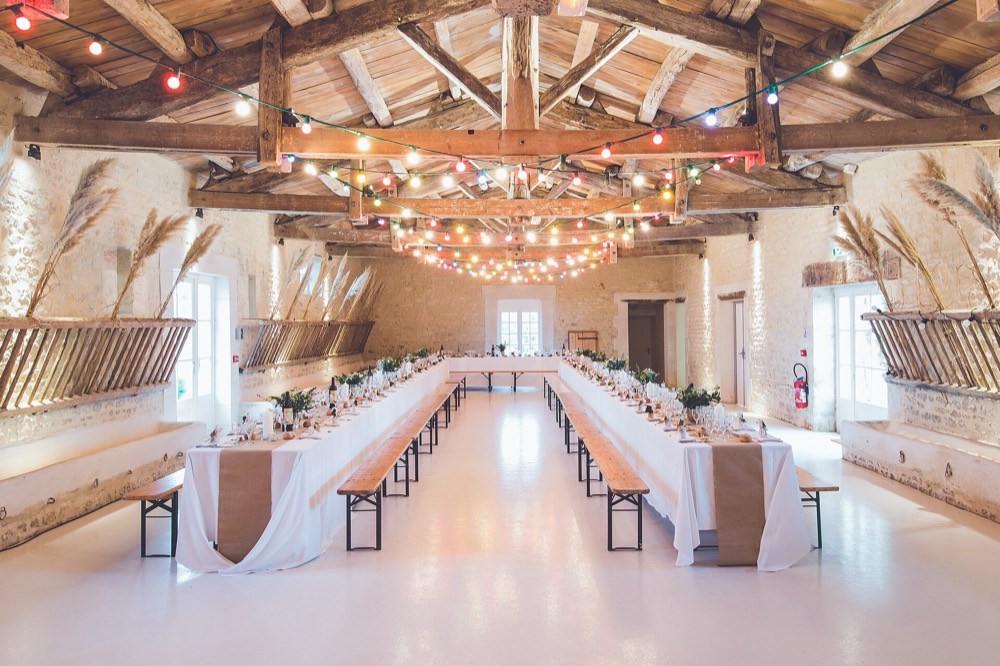 A long timber-trussed hall with stone walls, two rows of banquet tables and benches, and string lights across the beams
