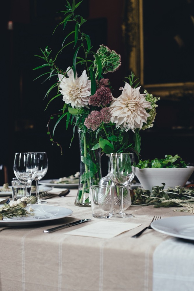 A dinner table set with linen, wine glasses and a vase of pale dahlias against a dark wall