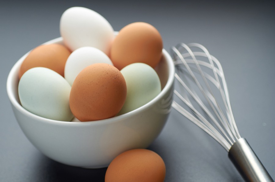 A white bowl of brown, white and pale green eggs beside a steel whisk